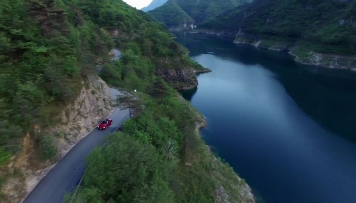 Fiat 124 Spider in movimento su strada che costeggia un lago di montagna.